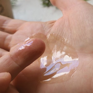 May include: A clear, viscous liquid is spread on a person's hand. The liquid is thick and has a slight sheen. The liquid is being spread with a finger.