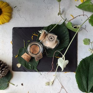 May include: Two small glass jars with cork lids and labels. The jars are filled with a light brown powder and are sitting on a black slate surface. The jars are surrounded by green leaves and flowers.