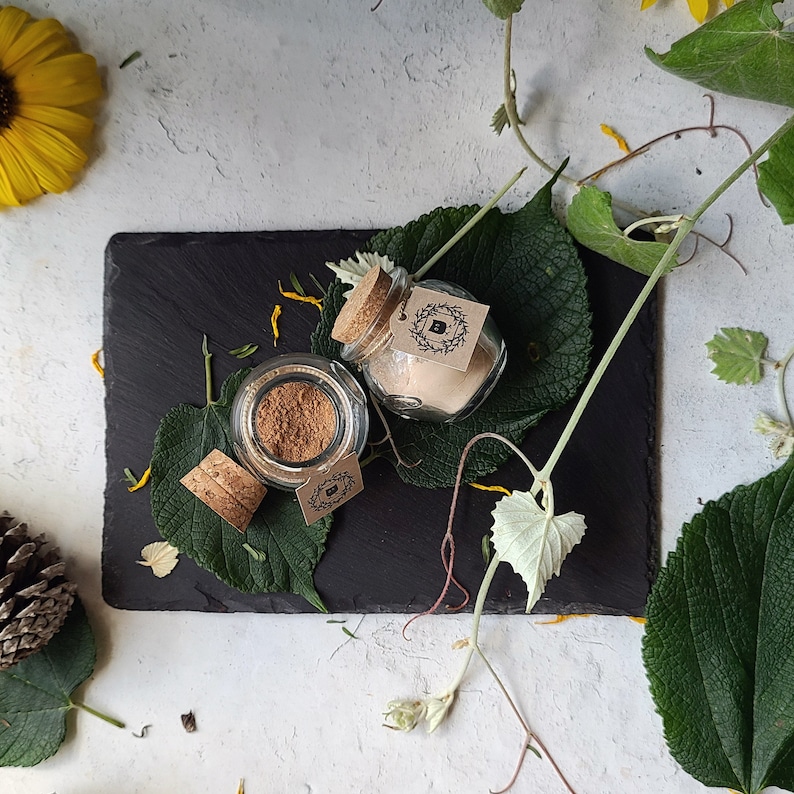 May include: Two small glass jars with cork lids and labels. The jars are filled with a light brown powder and are sitting on a black slate surface. The jars are surrounded by green leaves and flowers.