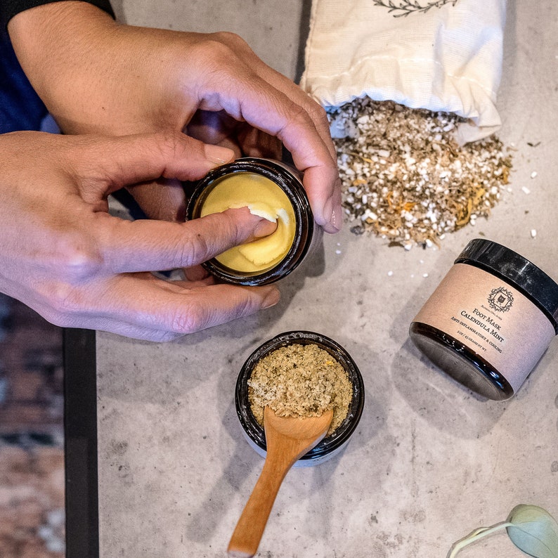 May include: Two small glass jars with lids, one containing a yellow balm and the other containing a green herbal foot mask. The foot mask jar has the text "Foot Mask Calendula Mint Anti-Inflammatory & Cooling" on the label.
