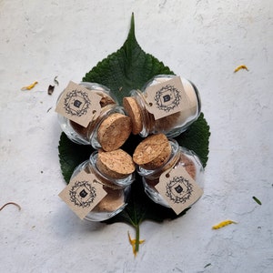 May include: Five small glass jars with cork lids and brown paper labels. Each label has a black and white illustration of a wreath with a letter inside. The jars are arranged in a circle on a white surface.
