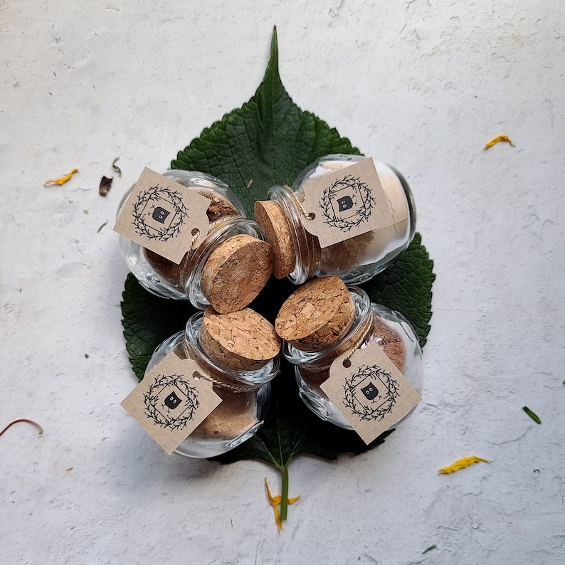 May include: Five small glass jars with cork lids and brown paper labels. Each label has a black and white illustration of a wreath with a letter inside. The jars are arranged in a circle on a white surface.