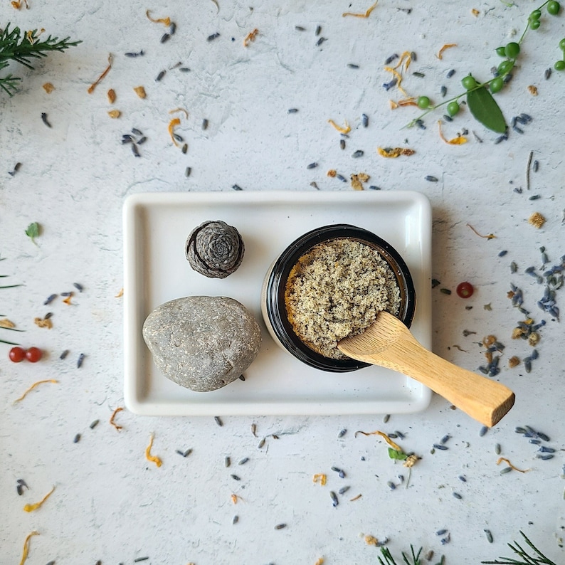 Pu&ograve; includere: Un vassoio in ceramica bianca con una piccola pigna, una pietra rotonda grigia, un piccolo barattolo di vetro pieno di uno scrub marrone chiaro e un cucchiaio di legno.