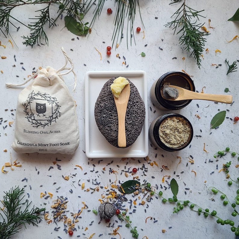 Foot Soak Bag, Lava Pumice, Scoop of Foot Balm on wooden spoon, Foot Mask jar top view with scoop of product on spoon, and top view of Foot Scrub jar. Scattered flowers and leaves.