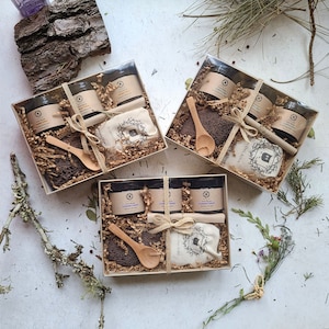 May include: Three gift boxes filled with bath and body products. Each box contains a variety of bath salts, soaps, and a wooden spoon. The boxes are decorated with natural elements like wood and twine.