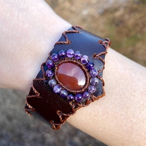 May include: A brown leather bracelet with a carnelian stone surrounded by amethyst beads. The bracelet is stitched with brown thread.