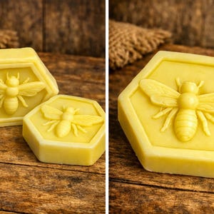 May include: Two hexagonal, yellow soap bars with a bee design. The bee is centered within the hexagon, with detailed wings and body. The soap bars are on a wooden surface, with a burlap sack in the background.