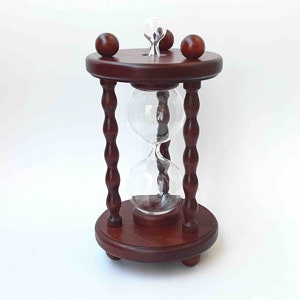 May include: A wooden hourglass with a dark brown finish. The hourglass has a round base and top with decorative wooden knobs. The glass bulbs are clear and contain a white sand-like substance.