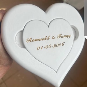 May include: A white heart-shaped wooden plaque featuring a smaller, inset heart piece. The center heart is engraved with the gold-colored text Romuald & Fanny and the date 01-08-2026. The design includes two circular cutouts on the sides.