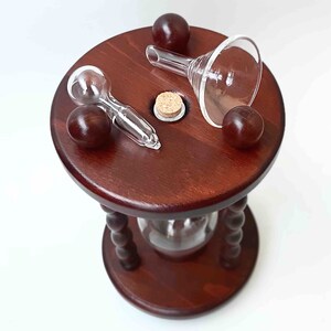 May include: A dark wood hourglass with a round top and base, featuring a clear glass funnel, spoon, and cork. Three dark brown spheres are placed around the top. The hourglass has a clear glass center and a decorative, turned wooden support.