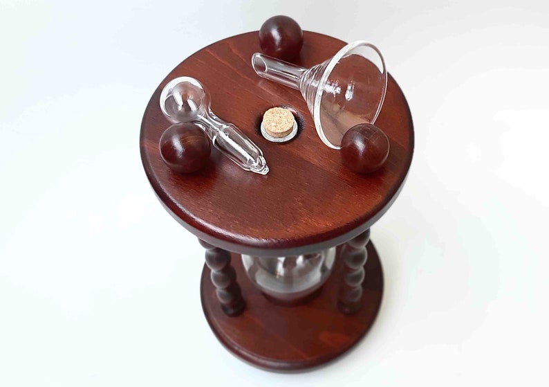 May include: A dark wood hourglass with a round top and base, featuring a clear glass funnel, spoon, and cork. Three dark brown spheres are placed around the top. The hourglass has a clear glass center and a decorative, turned wooden support.