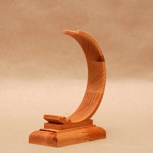 Puede incluir: Un soporte de madera para un reloj o una pulsera. El soporte tiene forma de media luna y un acabado liso y natural.