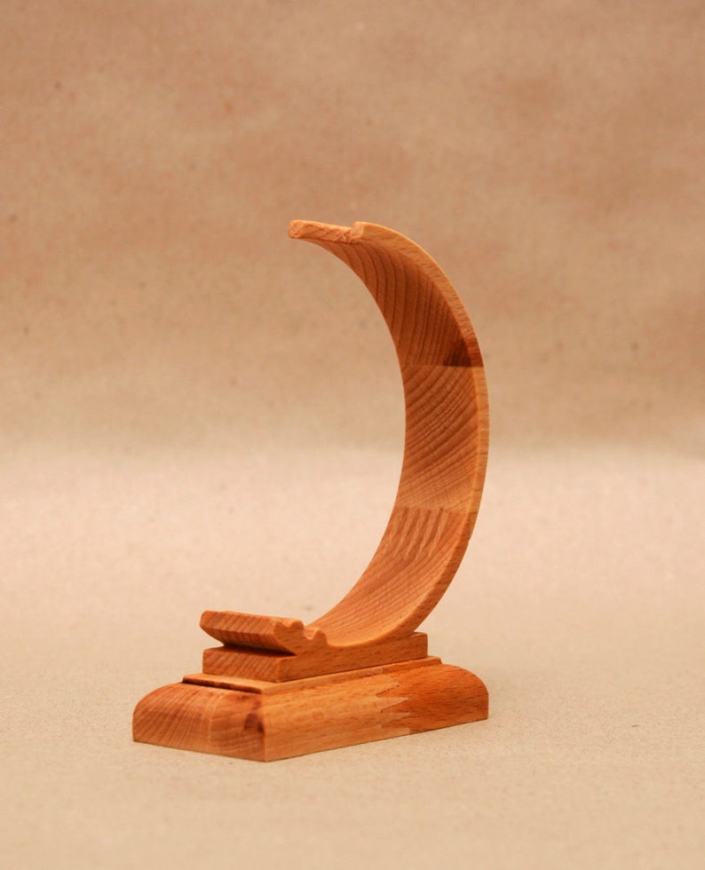 Puede incluir: Un soporte de madera para un reloj o una pulsera. El soporte tiene forma de media luna y un acabado liso y natural.