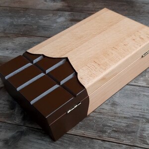 Puede incluir: Una caja de madera con un dise&ntilde;o de barra de chocolate. La caja est&aacute; hecha de madera clara y tiene un dise&ntilde;o de barra de chocolate marr&oacute;n oscuro en la parte delantera. La caja tiene una tapa con bisagras y est&aacute; cerrada.
