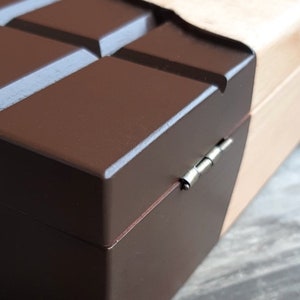 Puede incluir: Una caja de madera con forma de barra de chocolate con una tapa marr&oacute;n y una base de madera clara. La caja tiene una bisagra plateada.