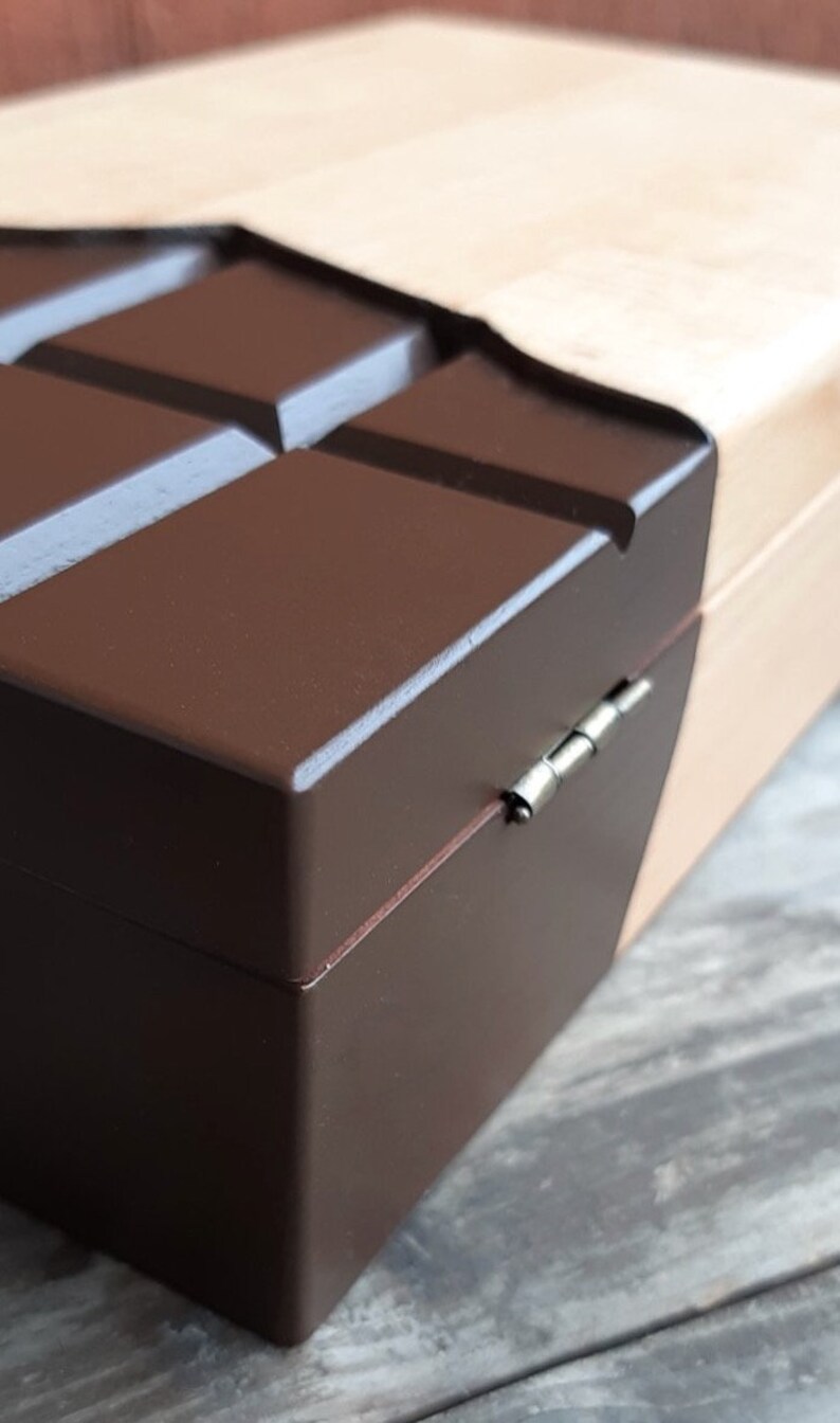 Puede incluir: Una caja de madera con forma de barra de chocolate con una tapa marr&oacute;n y una base de madera clara. La caja tiene una bisagra plateada.