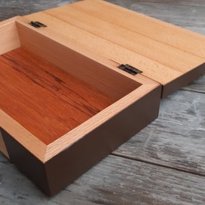 Puede incluir: Una caja de madera con tapa con bisagras, la caja est&aacute; te&ntilde;ida de un color marr&oacute;n oscuro y la tapa es de color madera natural. La caja est&aacute; abierta y el interior est&aacute; forrado con madera marr&oacute;n rojiza.
