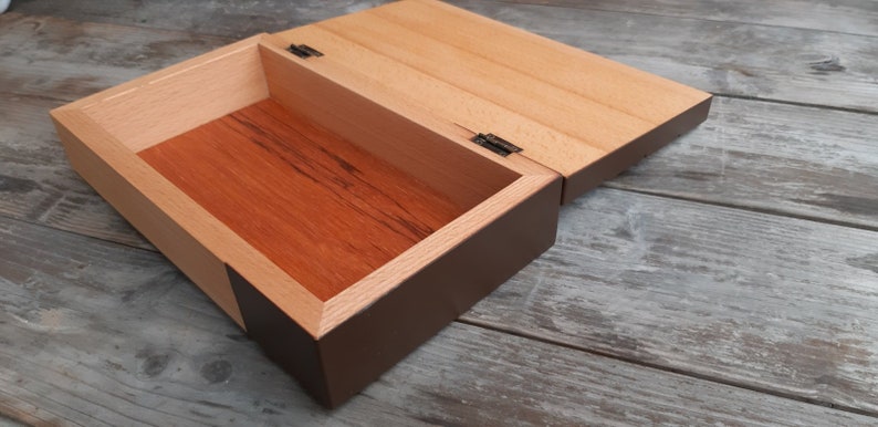 Puede incluir: Una caja de madera con tapa con bisagras, la caja est&aacute; te&ntilde;ida de un color marr&oacute;n oscuro y la tapa es de color madera natural. La caja est&aacute; abierta y el interior est&aacute; forrado con madera marr&oacute;n rojiza.
