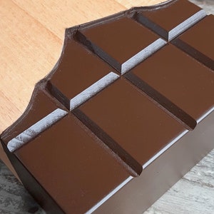 Puede incluir: Una tabla de cortar de madera con forma de barra de chocolate. La tabla es de madera clara y tiene un dise&ntilde;o de chocolate marr&oacute;n oscuro.