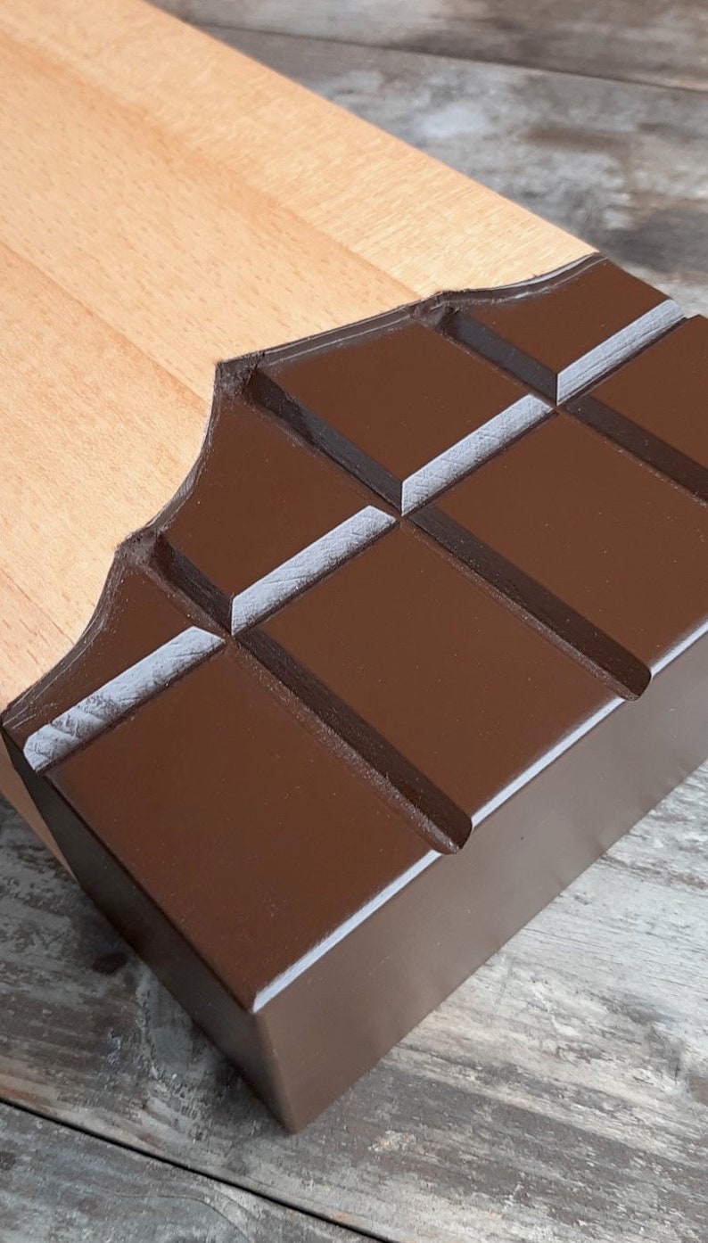 Puede incluir: Una tabla de cortar de madera con forma de barra de chocolate. La tabla es de madera clara y tiene un dise&ntilde;o de chocolate marr&oacute;n oscuro.