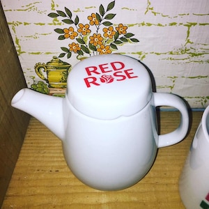 Peut inclure: Théière en céramique blanche avec un couvercle rouge sur lequel est inscrit "Red Rose" en lettres rouges. La théière est posée sur une surface en bois.