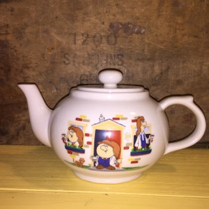 May include: A white ceramic teapot featuring a colourful cartoon illustration of three characters in a house setting. The design includes a central doorway with a person in a cap, flanked by two windows with people holding cups, a small cat, and a bird.