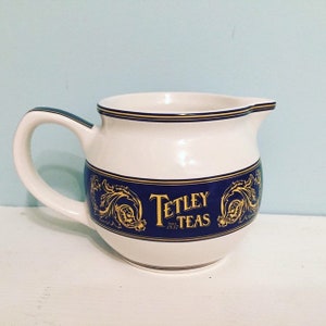 May include: White ceramic creamer with a blue and gold band featuring the text "Tetley Teas" and a gold floral design.