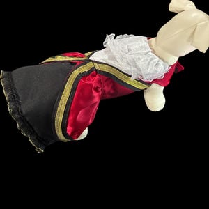 May include: A dog costume in the style of a royal outfit. The costume features a red satin top with gold trim, black pants with gold trim, and a white lace collar. The costume is designed to fit a dog.