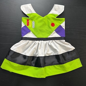 Puede incluir: Un vestido de disfraz inspirado en Buzz Lightyear. La parte superior es de color verde lima con detalles blancos, detalles morados y un círculo rojo. La falda tiene rayas blancas, negras y verde lima.