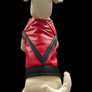 May include: A stuffed dog toy wearing a red and black vest. The vest has a V-shaped design on the back and a black band around the waist. The dog is cream-colored with floppy ears and a curled tail. The background is black.