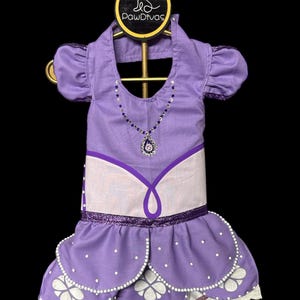 Puede incluir: Un vestido para mascota de color lavanda con mangas abullonadas, un collar falso con pedrería azul y transparente, y una falda de capas con detalles de perlas blancas y recortes florales. Incluye una etiqueta con el logo PawDivas y una tiara plateada.