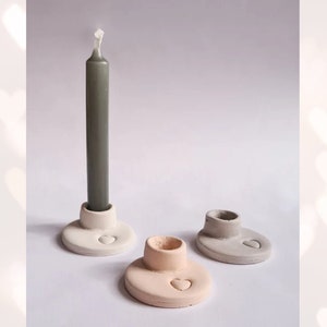 Peut inclure: Trois bougeoirs en céramique avec un motif en forme de cœur. Les bougeoirs sont en blanc, gris et rose clair. Un bougeoir contient une bougie grise.