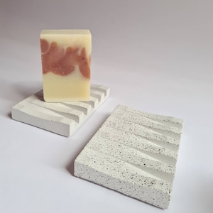 Puede incluir: Dos jaboneras de hormigón blanco con superficie texturizada. Una jabonera tiene un diseño en relieve con crestas y la otra tiene un diseño ondulado. Una pastilla de jabón con un patrón de remolino rosa y blanco está en la jabonera con crestas.