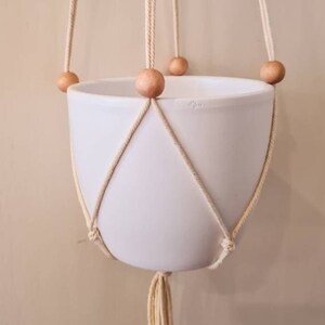 Op de afbeelding: Een witte keramische plantenpot die hangt aan een macrame plantenhanger met houten kralen. De hanger heeft een kwast aan de onderkant.
