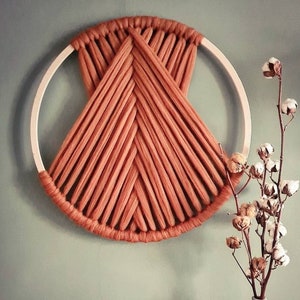 Op de afbeelding: Een macramé wandhanger met een geometrisch ontwerp in een warme oranje kleur. De hanger is gemaakt van dik garen en is bevestigd aan een houten ring. Een witte vaas met een katoenen stengelplant staat op de voorgrond.
