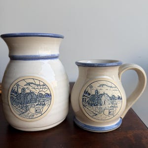 May include: A cream-colored ceramic pitcher and a matching mug, both featuring a blue rim and a circular decal depicting a farm scene with a barn, silo, and a map of the United States labeled MID AM. The items have a smooth, glossy finish.