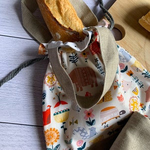 Puede incluir: Una bolsa de lona beige con un estampado floral y animal colorido. La bolsa tiene un cierre de cordón y está llena de una barra de pan.