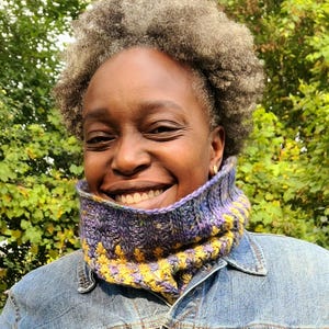 May include: A person wearing a blue denim jacket with a purple and yellow knitted neck warmer. The neck warmer has a textured pattern.