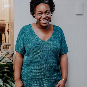 May include: A teal short-sleeved sweater with a V-neck. The sweater has a textured knit pattern and is paired with black pants. The person in the image is smiling and wearing earrings and a bracelet.
