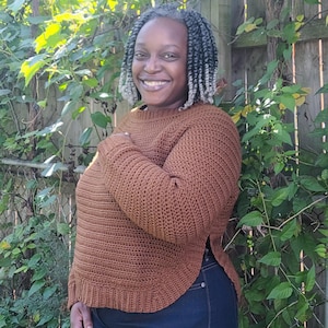 May include: A brown crochet sweater with long sleeves and a split hem. The sweater has a ribbed neckline and cuffs. The person is wearing dark blue jeans. The background includes a wooden fence and green foliage.