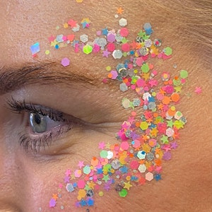May include: Close-up of a person's face with colourful glitter applied to the skin near the eye. The glitter is in the shape of stars and hexagons and is a mix of pink, blue, green, yellow, and white.