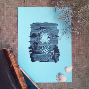 May include: A black and white linocut print on light blue paper depicts a serene moonlit scene with clouds, water, and a figure. The print is displayed with a vintage book and seashells, suggesting a coastal or nature theme.