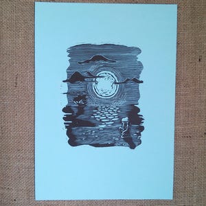 May include: A black and white linocut print on light blue paper. The artwork depicts a moonlit scene with a figure seated near water. The moon is surrounded by clouds, and the water reflects the moonlight. The artist's signature is visible.