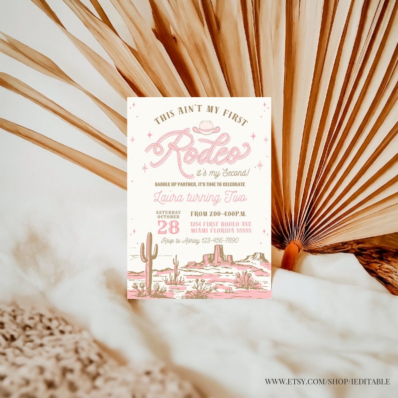 May include: A pink and brown invitation for a second birthday party with a western theme. The invitation says "This ain't my first Rodeo, it's my second!" and features a pink cowboy hat with a lasso. The invitation also includes the text "Saddle up partner, it's time to celebrate Laura turning two." The invitation is set against a background of a desert landscape with cacti and mountains.