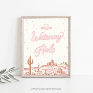 Könnte beinhalten: Ein druckbares Schild mit einem rosafarbenen Cowboyhut und dem Text "Watering Hole" in rosafarbener Schrift. Der Hintergrund ist eine weiße Wüstenlandschaft mit Kakteen und Bergen in Braun und Rosa.