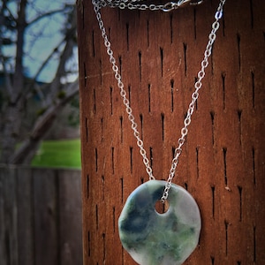 May include: A silver chain necklace with a round, green and gray pendant. The pendant has a hole in the center and is suspended from a delicate chain. The necklace is displayed against a brown wooden background.