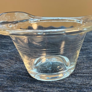 Könnte beinhalten: Eine klare Glasschale mit einem breiten, ausgestellten Rand und einem glatten, abgerundeten Boden. Die Außenseite der Schale weist zwei horizontale, eingravierte Linien auf, die den Mittelteil umkreisen und der transparenten Oberfläche Struktur verleihen.