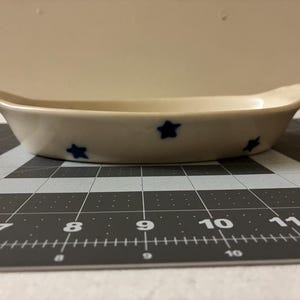 以下が含まれることがあります： ボートのような形をしたオフホワイトのセラミック製サービングディッシュ。ディッシュには、濃い青色の星のデザインが均等に配置されています。ディッシュは、黒とグレーのカッティングマットの上に置かれています。