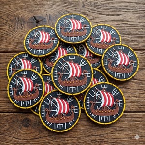 Op de afbeelding: Een verzameling ronde Viking schild patches met een gele rand. Elke patch toont een bruin Vikingschip met een rood-wit gestreept zeil. De patches liggen op een houten oppervlak.