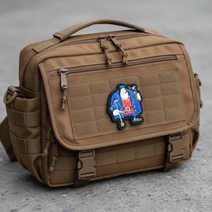 Puede incluir: Bolso mensajero marrón con múltiples compartimentos, cremalleras y correas ajustables. Un parche de dibujos animados de un hombre con abrigo azul y chaleco rojo está adherido en la parte delantera. El bolso tiene un asa superior y bolsillos laterales.